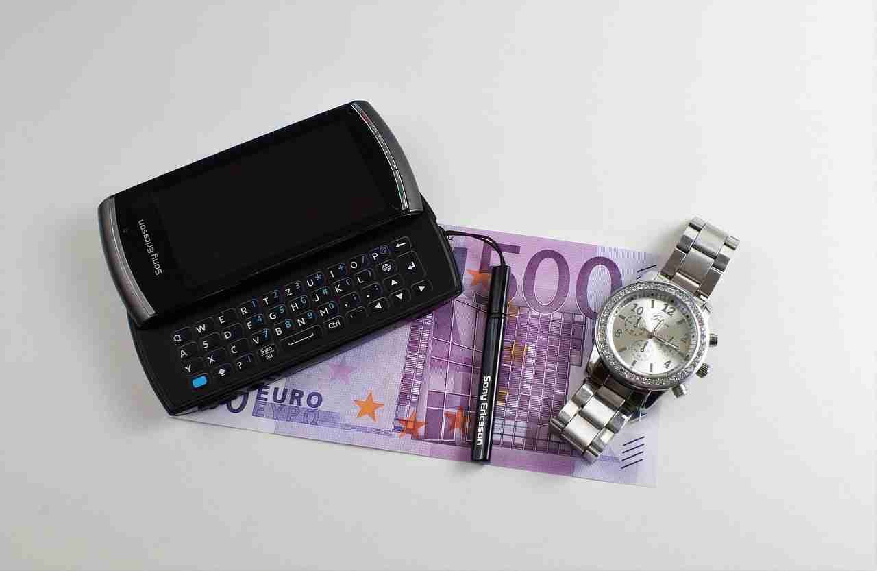 A smartphone with a sliding keyboard, a 100 euro banknote, and a stainless steel wristwatch laid out on a white surface.