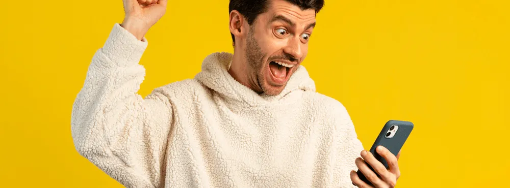 Ecstatic man in a white hoodie celebrating good news on his mobile phone against a bright yellow background. mobile phones bad credit.