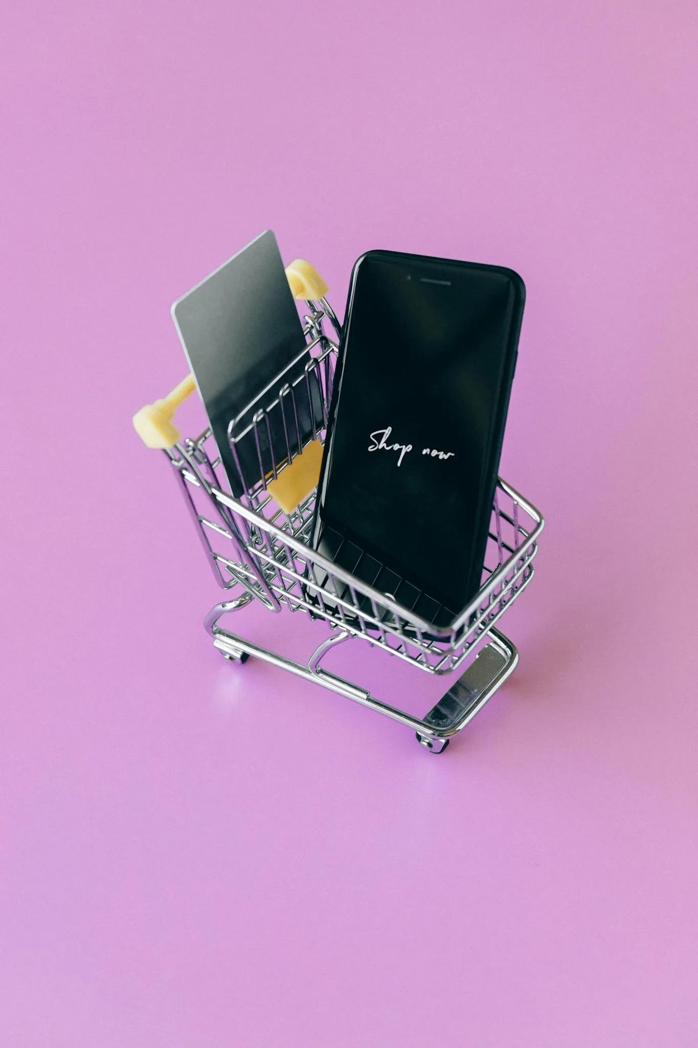 Modern shopping concept featuring smartphone and credit cards in a miniature cart on pink background.